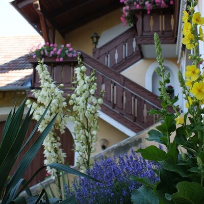 Die Fassade des Bauernhofs mit einer Holztreppe, blumengeschmückten Balkonen und vielfältiger Bepflanzung im Garten.