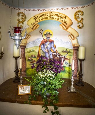 Ein kleiner Schrein auf dem Hof mit einem Wandgemälde des Heiligen Leonhard, geschmückt mit Blumen und Kerzen.
