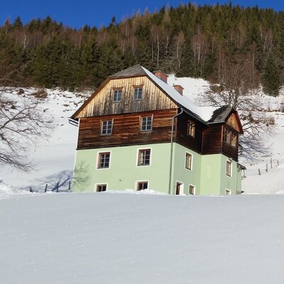 Der Hof mit seinen hellgrünen Wänden und Holzfassade, umgeben von schneebedeckter Landschaft und einem Wald im Hintergrund.