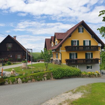 Die Gebäude des Bauernhofs, inklusive eines gelben Hauses mit Balkonen und eines dunklen Holzgebäudes, sind von Gärten und Weinbergen umgeben.