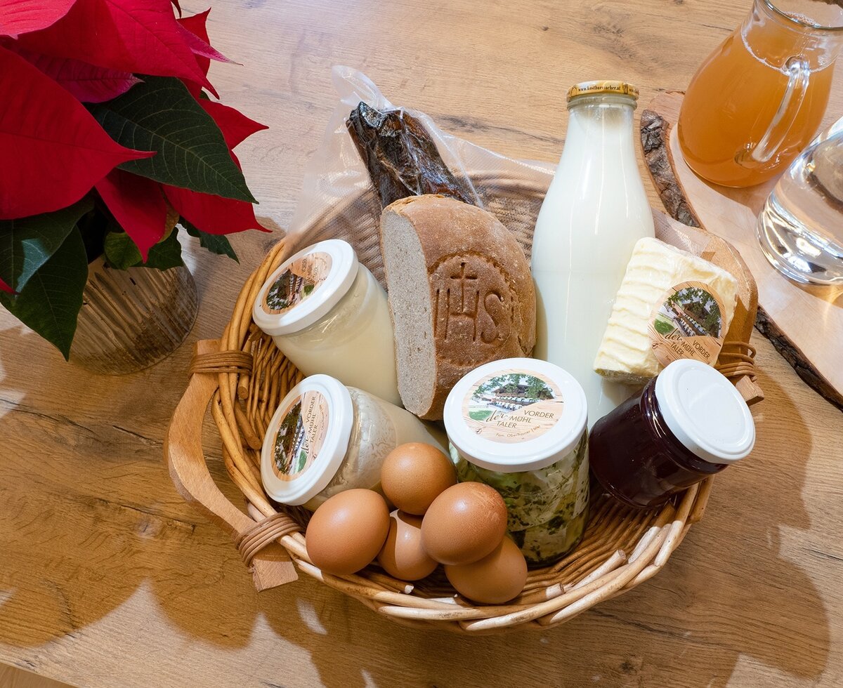 Ein Korb gefüllt mit frischen Produkten vom Bauernhof, darunter Milch, Brot, Butter, Eier, Konfitüre und Aufstriche.